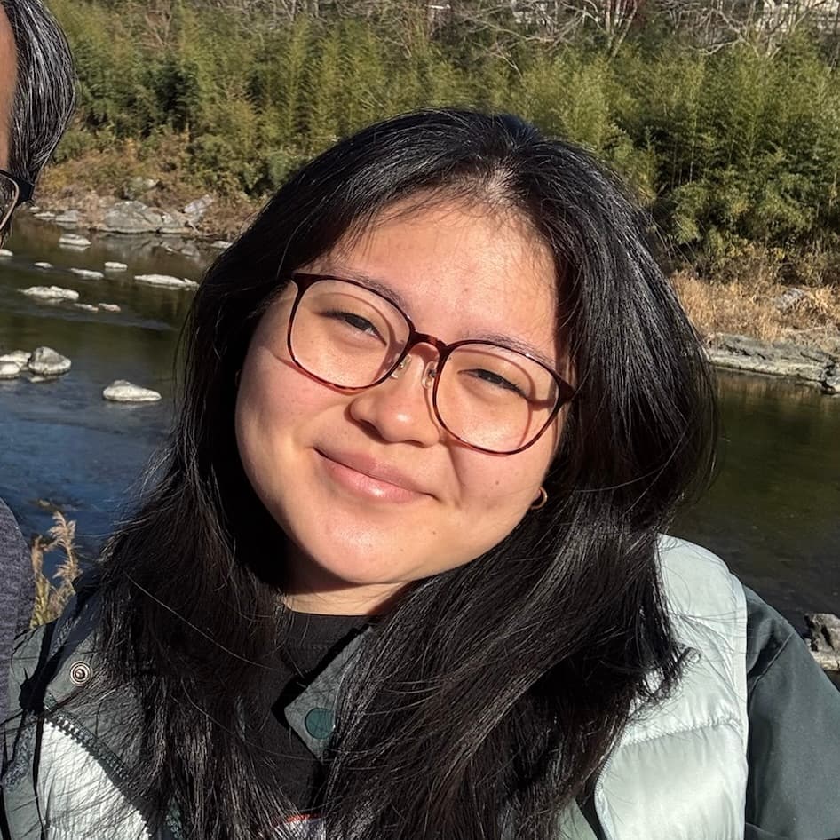 Selfie of Reina smiling. They are wearing brown square-ish glasses and long black hair.